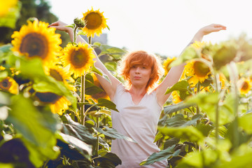 young girl enjoying nature on the field of sunflowers at sunset, portrait of the beautiful redheaded woman girl with a sunflowers in a sunny summer evening, hands raised up