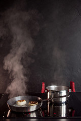 Frying pan and cooking pot on induction hob, steam rises. Black textured kitchen