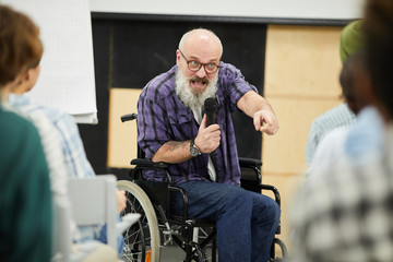 Fototapeta na wymiar Emotional confident motivational disabled speaker with gray beard sitting in wheelchair and pointing at person in audience while helping students to believe in themselves at conference.