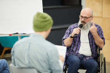 Serious confident senior bald man with beard sitting in wheelchair and speaking into microphone while holding presentation for students at conference