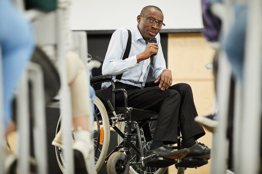 Serious Confident Motivational Disabled Mature African-American Speaker In Glasses Sitting In Wheelchair And Looking At Audience While Saying Speech Into Microphone At Conference