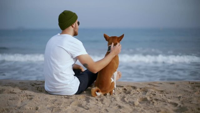 Young hipster man in green beanie and white tshirt with authentic arm tattoo relaxes on beach with best friend puppy or brown dog, looks away to horizon with smile, pets animal
