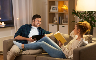 leisure and people concept - happy couple with tablet computer and book talking at home in evening