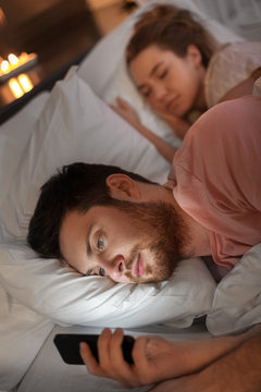 technology, internet addiction and cheat concept - man using smartphone at night while his girlfriend is sleeping