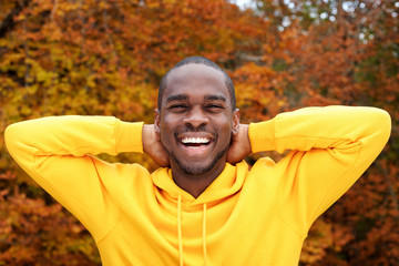 handsome young african american man smiling with autumn leaves in background and hands behind head