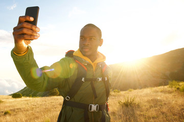 young black man hiking in mountains taking selfie with mobile phone and sunset in background