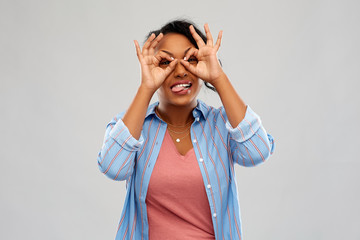 people, gesture and fun concept - happy african american young woman looking through finger glasses and sticking tongue out over grey background