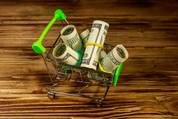 Small shopping cart with rolls of one hundred dollar bills on wooden background. Consuming concept