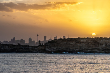 Fototapeta premium Sydney, Australia - February 12, 2019: Sunset over city skyline seen from Tasman Sea. Shoreline rocky cliffs. Yellow brown sky, sun rays. 3 of 5.