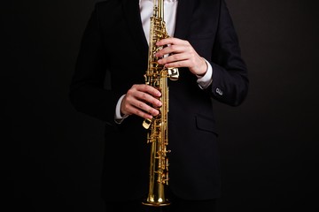 Male saxophonist in a black classic suit playing on the soprano saxophone while standing on a black...