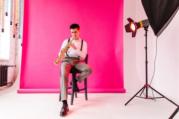 Saxophonist in classic clothes playing the soprano saxophone sitting on a chair on a pink background in a photo Studio