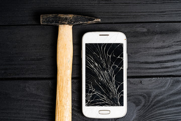 Smartphone smashed by hammer isolated on black wooden background. Broken touchscreen of a smartphone lying close to the hammer on table.