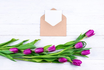 Composition of white blank card in opened craft envelope decorated with fresh beautiful purple tulips on white wooden background with copy space.