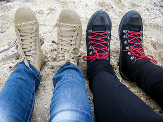 Female legs girlfriends on the beach in shoes. Gray and black sneakers with red laces in jeans. Sand by the sea. Lifestyle travel.