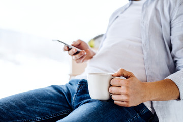 Fototapeta na wymiar A midsection of man with smartphone and coffee sitting indoors, text messaging.