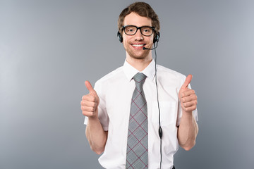 smiling call center operator in glasses and headset showing thumbs up on grey background
