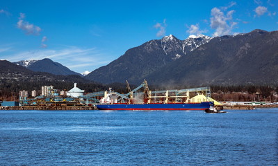 Terminal of North Vancouver port on a background of mountain scenery. The vessel is under load, the tug is towing another vessel.