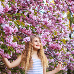 Adorable little girl with curly hair in pink dress walking in blossom cherry garden in sunny spring day