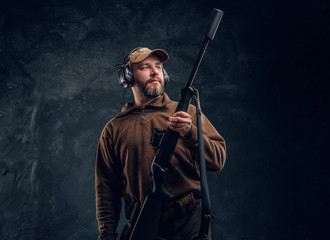 Portrait of a hunter in cap and headphones holding a rifle and looking sideways. Studio photo against a dark wall background