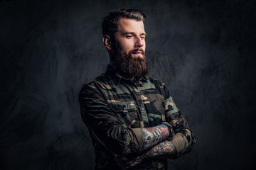 A stylish bearded guy with tattooed hands in the military shirt, posing with his arms crossed and looking sideways. Studio photo against dark wall