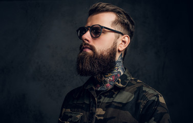 Closeup portrait of a bearded man with a tattoo on his neck in sunglasses wearing a military shirt. Studio photo against dark wall