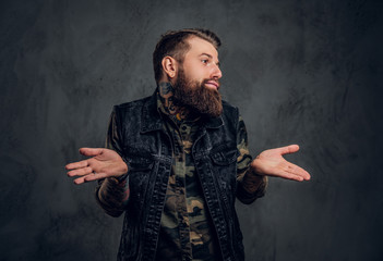 A stylish bearded guy with tattooed hands in military shirt and denim vest posing with a puzzled look. Studio photo against dark wall