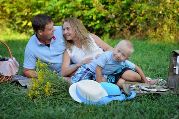 Fototapeta premium Family picnic in nature. Mom, dad and son relax sitting on the grass in the Park in the summer at sunset. A little boy looks at the camera and smiles. Portrait. Horizontal orientation of the image