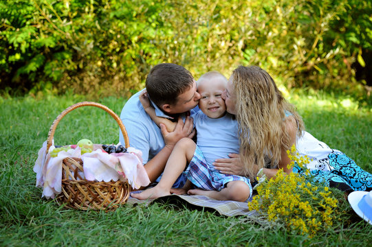 Mom and dad together kiss smiling little son sitting on the grass in the Park in the summer at sunset. Family picnic outdoors. Portrait. Horizontal orientation of the image