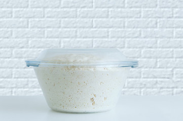 Yeast dough made from wheat flour. In a glass dish on a white table. Background brick wall.