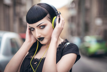Urban goth girl listening to her favourite music over her big headphones, street in a city surroundings