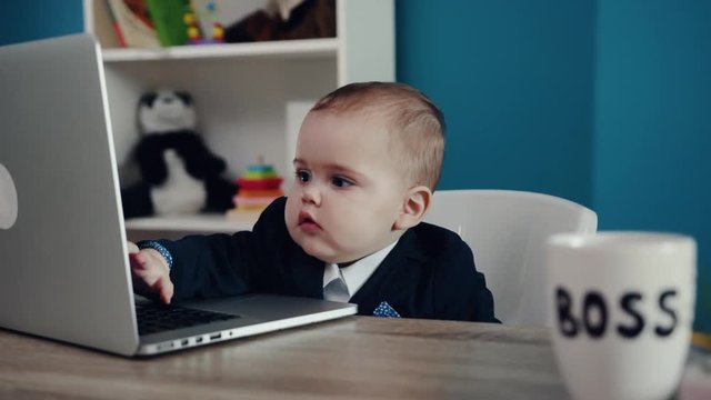 Photo of adorable serious baby boss in business suit typing on keyboard of the laptop while sitting in the modern office. Success, happy childhood, cuteness. Child�s portrait. Close up view