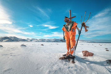 Scarecrow in orange clothes and gas mask with trident is against blue sky with clouds and snowy...