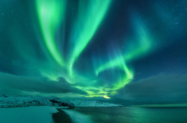 Fototapeta na wymiar Aurora borealis over ocean. Northern lights in Teriberka, Russia. Starry sky with polar lights and clouds. Night winter landscape with aurora, sea with stones in blurred water, snowy mountains. Travel