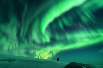 Aurora and silhouette of alone standing man on the hill. Lofoten islands, Norway. Aurora borealis and photographer. Sky with stars and green polar lights. Night landscape with northern lights. Travel