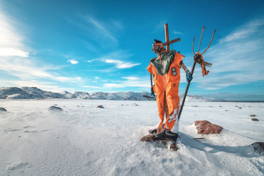 Scarecrow in orange clothes and gas mask with trident is against blue sky with clouds and snowy meadow. Winter landscape with funny dummy, stones in snow and mountains in Teriberka, Russia. Travel 