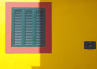 a vibrant yellow wall with red frame and green closed shuttered window typical Portuguese colors in bright sunlight and shade with space for copy