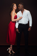 beautiful woman in red dress looking at camera and undressing african american man isolated on black