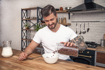 young happy handsome smiling guy is preparing his breakfast at the kitchen mixing cereal with milk