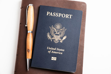 The iconic dark blue cover of an American passport with brown leather-covered journal and a ballpoint pen held together with an elastic band artistically set on a plain white background.