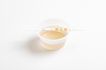 A set of urine sample on a round plastic cup with a single unused urine reagent strip against a plain white background.