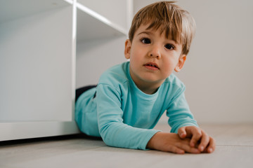 Naklejka premium Portrait of a little boy in blue at home lying in the book shelf on the floor hiding games