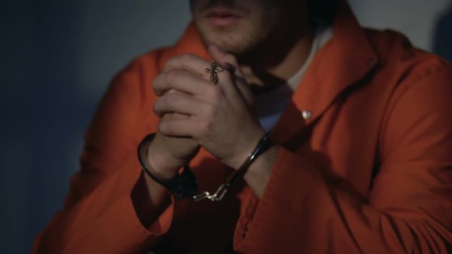 Imprisoned male in handcuffs holding cross and praying, feeling guilty, regret