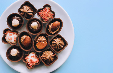 Beautiful chocolates of different shapes and fillings lie in a white plate against the blue background paper. Copy space, top view.