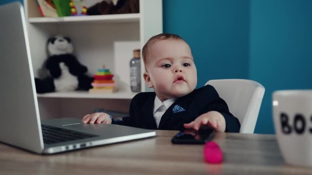 Close up view of extremely cute baby boss in formal suit sitting by the laptop and taking the phone of the table. Toddler shooting, indoors. Modern kids office. Success, happy childhood, cuteness