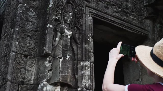 Woman tourist is taking picture of deity carvings on the stone wall using smartphone. Bayon Temple, a richly decorated Khmer temple at Angkor in Cambodia built in the late 12th century