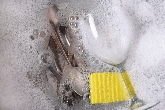 Dishpan filled with soapy water with cutlery, a wine glass and sponge
