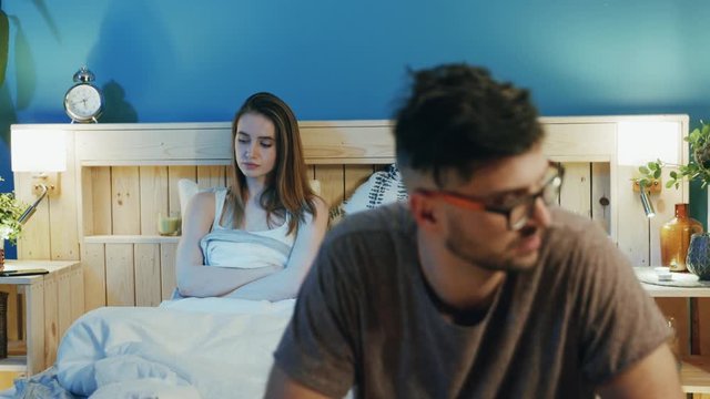 Burred image of young man in glasses sadly sitting on the edge of bed, focus on disappointed woman. Conflict situation, stress, bad mood, negative emotions. Problems in family life, troubles
