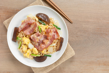 top view of stir fried instant noodle with bacon and vegetable in a ceramic dish on wooden table. asian homemade stlye food concept.