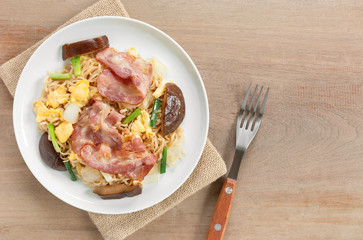 top view of stir fried instant noodle with bacon and vegetable in a ceramic dish on wooden table. asian homemade stlye food concept.