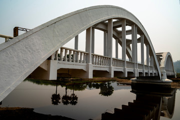 Side view of concrete white railroad bridge with reflection in the river and blue sky background.